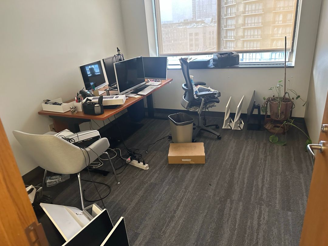 A private office with a desk, monitors and chairs beside a high-rise window, before the cleanout.