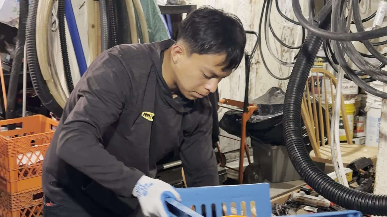 A Junk B Gone crew member sorting tools and extension cords into a blue crate in a packed basement workshop
