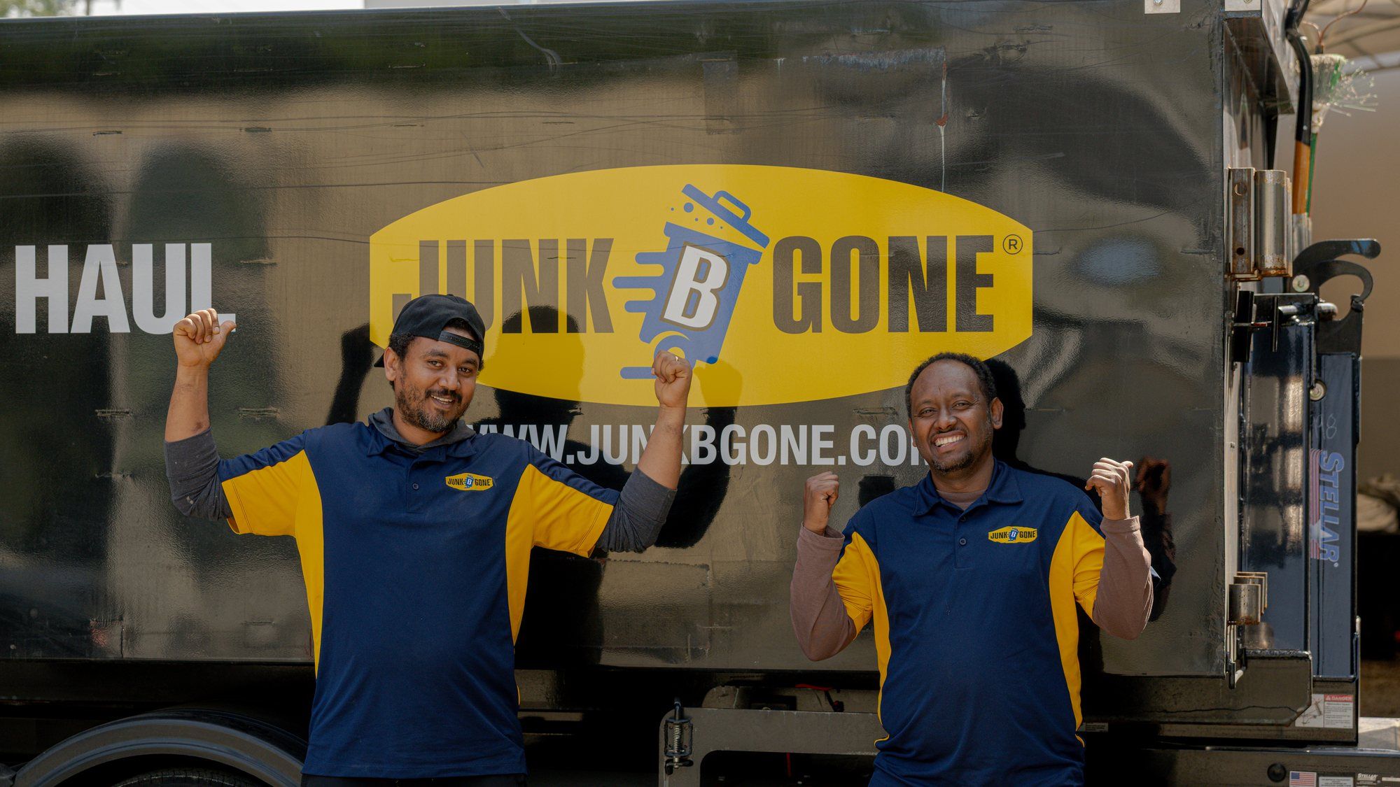 Two Junk B Gone crew members in company polos cheer in front of a company truck.