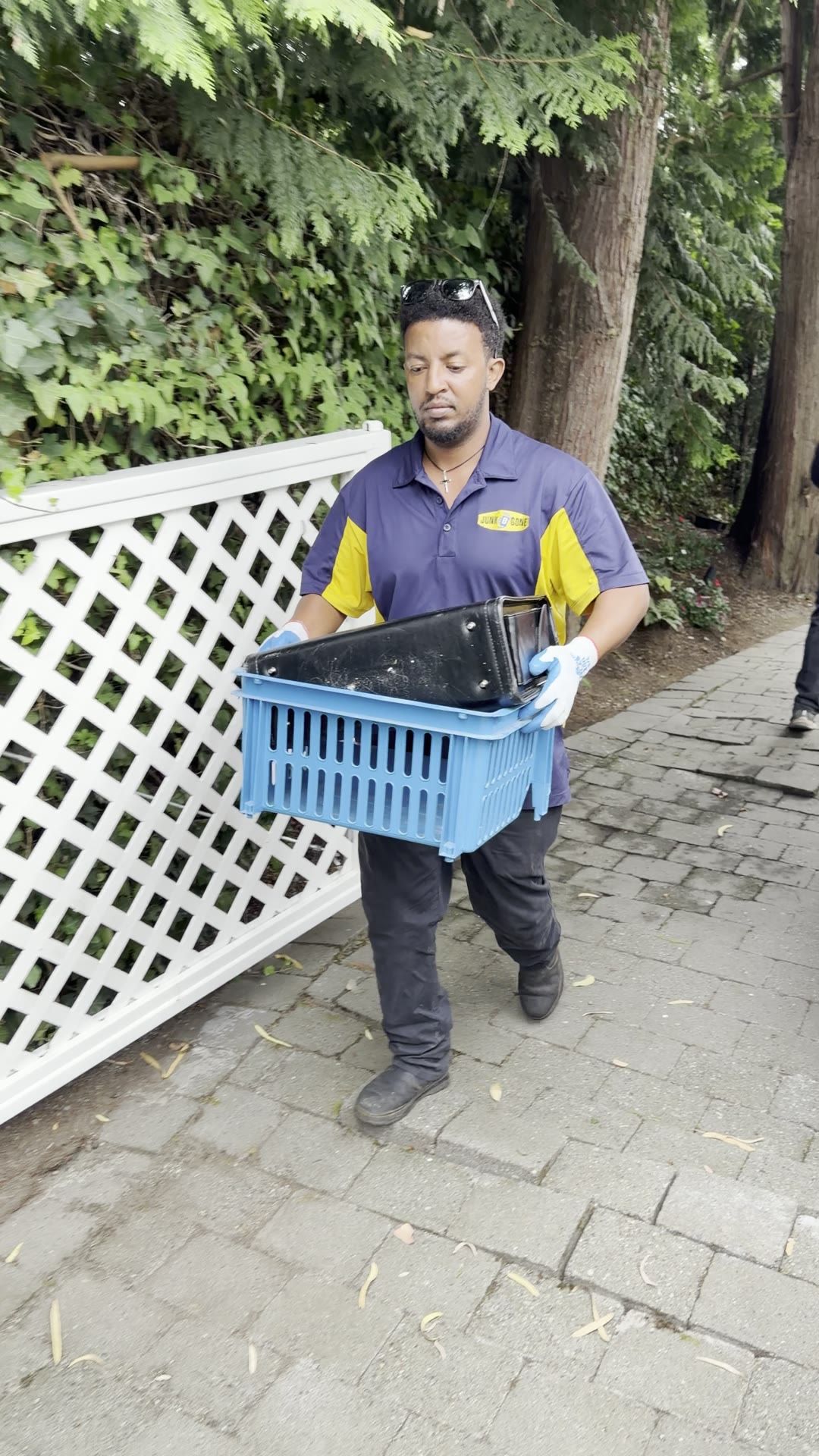 A Junk B Gone crew member in a company polo carries a blue crate along a garden path.