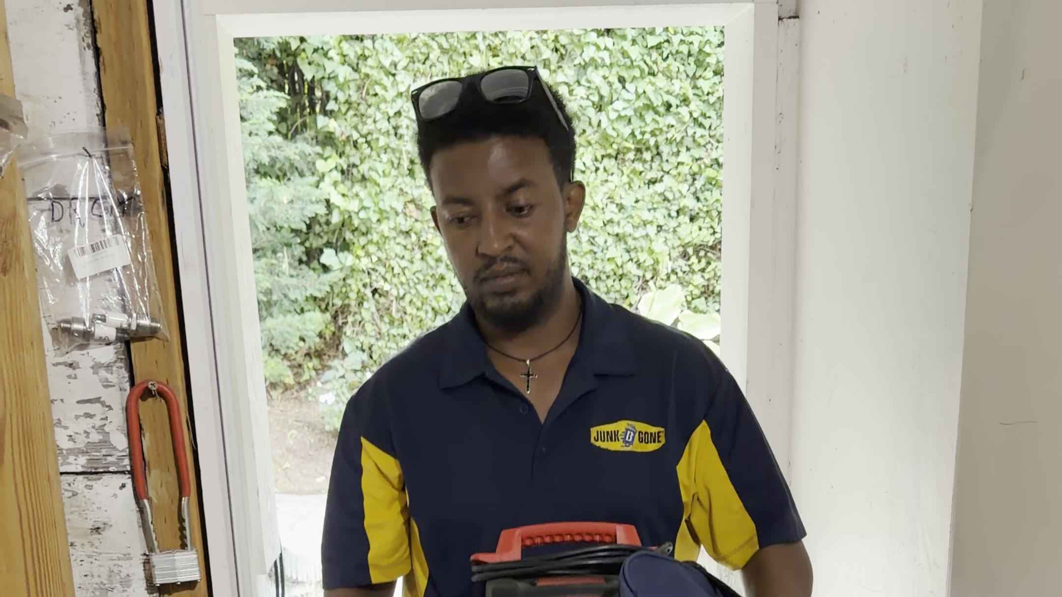 A Junk B Gone crew member in a company polo carries a shop vac and a bag through a doorway.