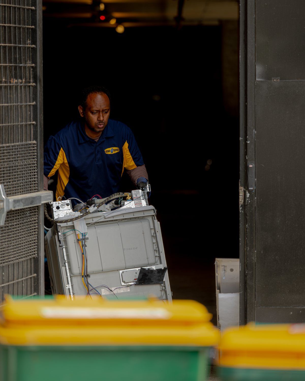 A Junk B Gone crew member in a company polo wheels an appliance out through a building service door past the recycling bins.
