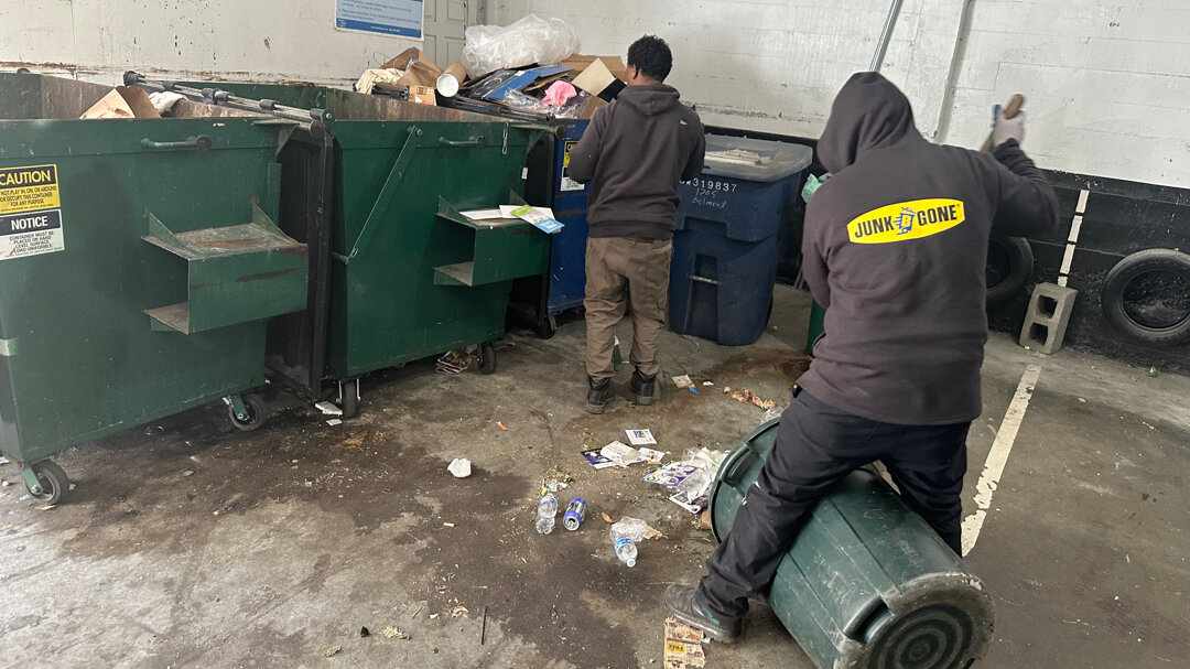 A Junk B Gone crew member in a branded hoodie clears an overflowing trash room beside full dumpsters.