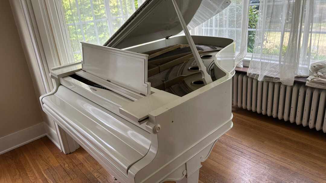 A white grand piano beside a bay window in a bright living room with hardwood floors.