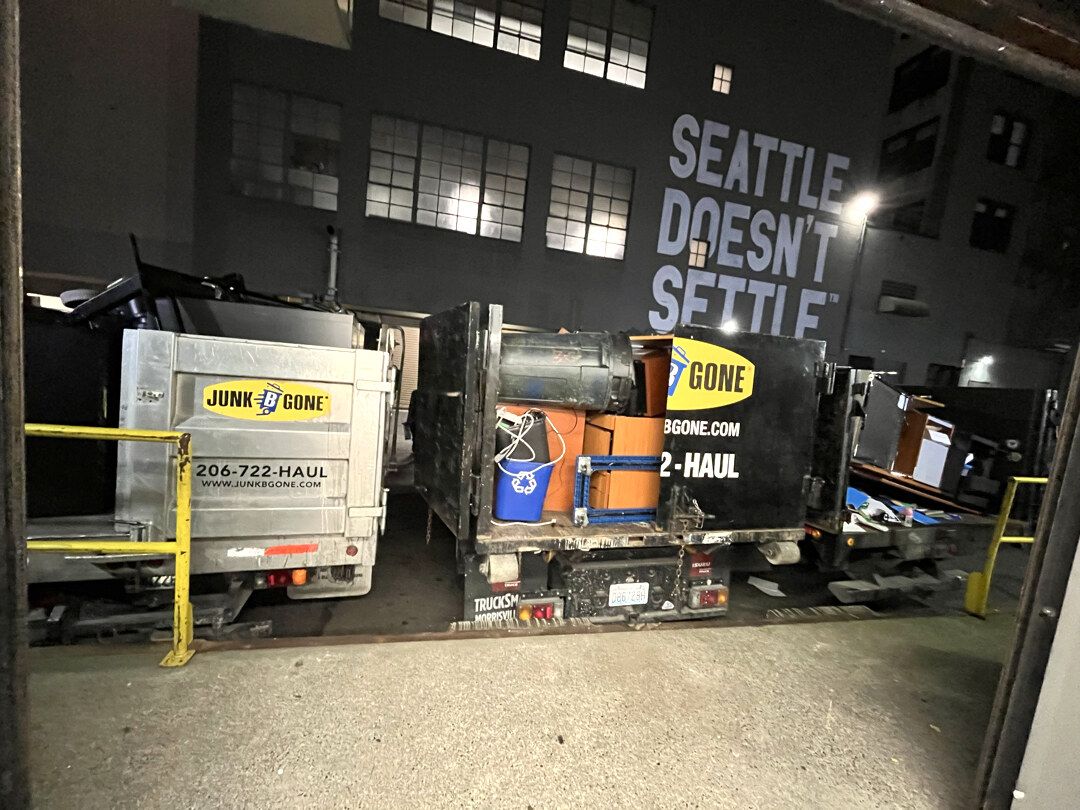 Three Junk B Gone trucks parked side by side at a loading dock at night, beds loaded with office furniture, boxes and a blue recycling bin, under a building mural that reads Seattle Doesn't Settle.